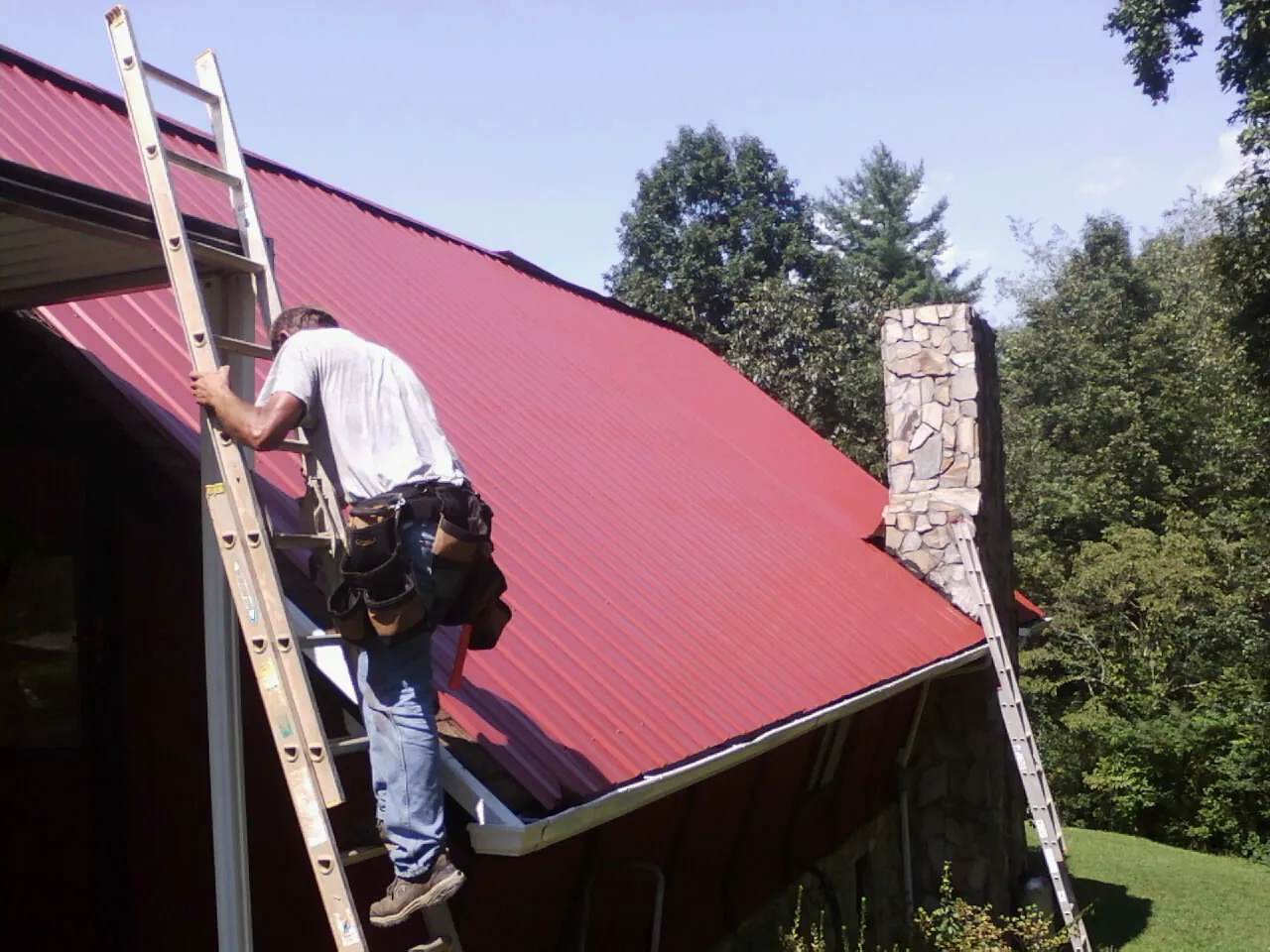 Roofer on a ladder beside a red metal roof and stone chimney — Soffit & Fascia Repair by Roofing Brothers in Fairchild Air Force Base